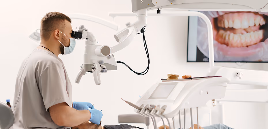 Dentist wearing gloves and mask examines patient’s teeth using a dental microscope with teeth image on screen.