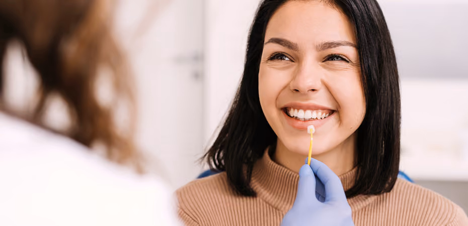 Smiling woman being examined with a dental shade guide by a gloved hand in a clinic.
