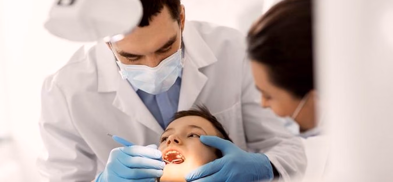 Dentist wearing gloves and mask examining a young boy's teeth while a woman watches closely.
