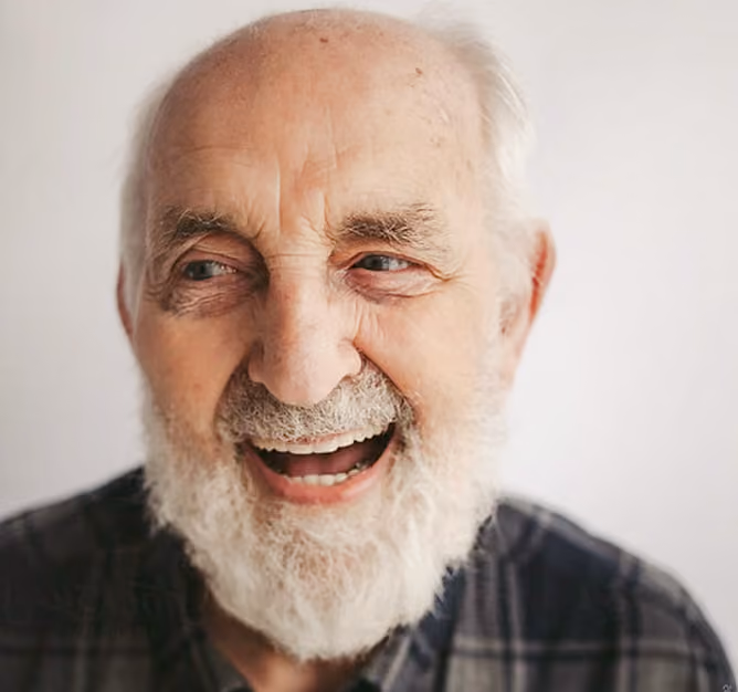 Smiling elderly man with white beard wearing a plaid shirt against a light background.